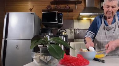 Modern bir ev mutfağında yemek hazırlayan kısa gri saçlı yaşlı bir kadın. İçeriğiyle dikkatlice ilgilenir, ev ortamında deneyimli bir şekilde yemek pişirme sürecine odaklanır. 