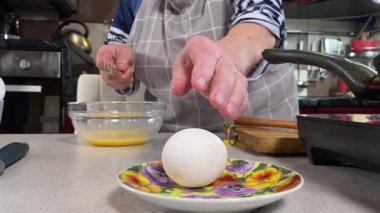 Modern bir ev mutfağında yemek hazırlayan kısa gri saçlı yaşlı bir kadın. İçeriğiyle dikkatlice ilgilenir, ev ortamında deneyimli bir şekilde yemek pişirme sürecine odaklanır. 