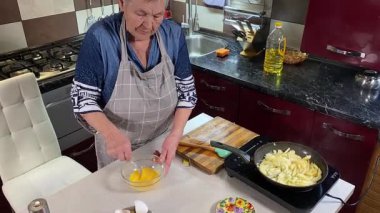 Modern bir ev mutfağında yemek hazırlayan kısa gri saçlı yaşlı bir kadın. İçeriğiyle dikkatlice ilgilenir, ev ortamında deneyimli bir şekilde yemek pişirme sürecine odaklanır. 