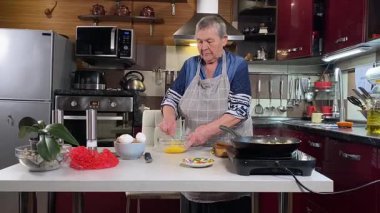 Modern bir ev mutfağında yemek hazırlayan kısa gri saçlı yaşlı bir kadın. İçeriğiyle dikkatlice ilgilenir, ev ortamında deneyimli bir şekilde yemek pişirme sürecine odaklanır. 