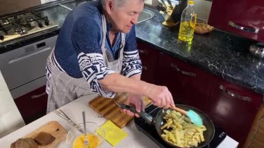 Modern bir ev mutfağında yemek hazırlayan kısa gri saçlı yaşlı bir kadın. İçeriğiyle dikkatlice ilgilenir, ev ortamında deneyimli bir şekilde yemek pişirme sürecine odaklanır. 