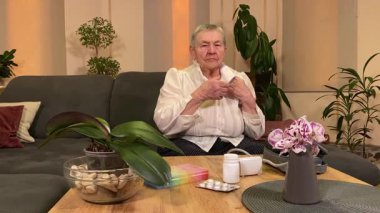 Yaşlı bir kadın, sıcaklığı, oturma odasındaki kan basıncını ölçerek sağlıkla ilgilenir. Günlük ilaç organizatörü ve ilaç çevrede görünüyor, rutin ve tıbbi bakımını yansıtıyor. 