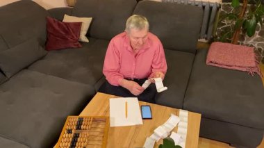 Oturma odasında vergileri, elektrik faturalarını akıllı telefon ve abaküs kullanarak hesap yapan yaşlı bir kadın. Odaklanmış ve düşünceli. Ekonomisini hem modern hem de geleneksel araçlarla yönetiyor. 