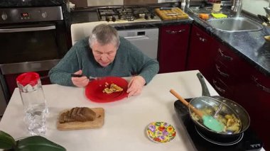 Sıcak bir ev mutfağında öğle yemeği yiyen, ev yapımı sıcak yemeğin tadını çıkaran yaşlı bir kadın. Yavaş ve dikkatli bir şekilde yiyor, tanıdık yiyeceklerin rahatlığını ve etrafındaki sakin atmosferi takdir ediyor.. 