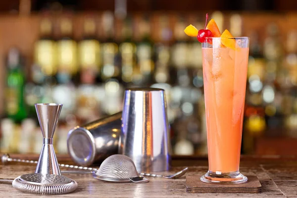 stock image Cocktail next to a shaker at a bar on a blurry background of bottles with alcohol in a nightclub or in a pub. Place for text.