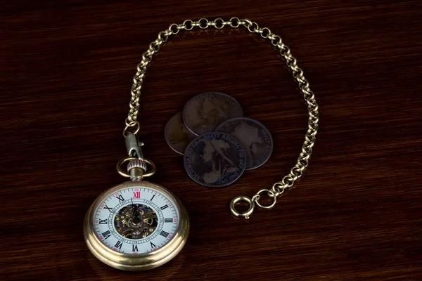 Old victorian UK pennies with retro pocket watch on a varnished wooden table top