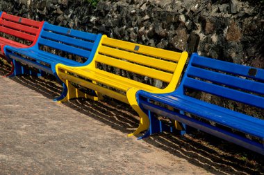 Sahildeki bir geçidin duvarında çeşitli renklerde kamusal banklar.