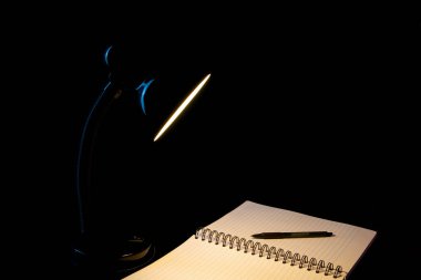 Açık çizgili bir not defteri ve tükenmez kalemle siyah bir arkaplanı aydınlatan siyah bir masa lambasının yakınlaştırılması