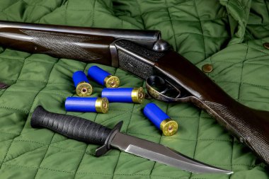 Örtülü bir ceketin üzerinde av bıçağı ve av tüfeği fişekleri olan bir av tüfeği.