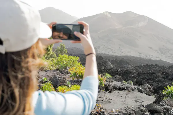 woman with a cap taking a horizontal photo of the Tajogaite volcano with her cell phone and she sees herself reflected in the phone, she comes out of focus in the foreground