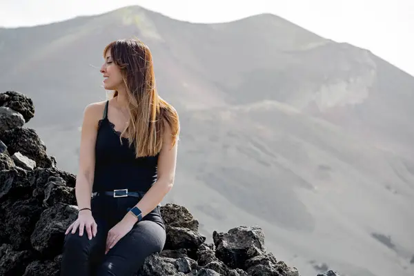 woman in suspenders looks at the ground smiling in front of the Tajogaite volcano, the volcano is seen out of focus in the background, at a viewpoint on the island of La Palma
