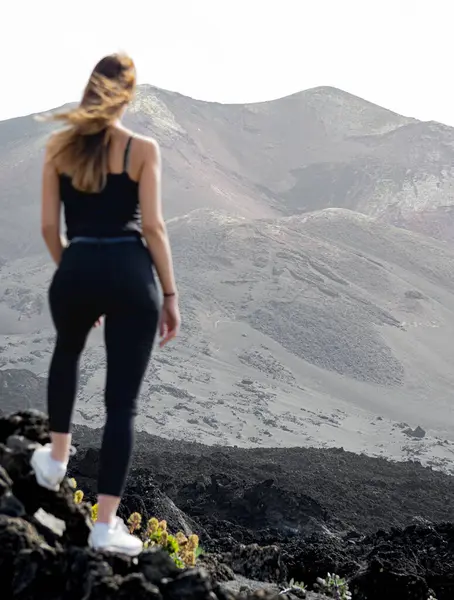 woman in suspenders standing with her hair flying in the wind looking at the Tajogaite volcano from a viewpoint, on top of some rocks, on the island of La Palma
