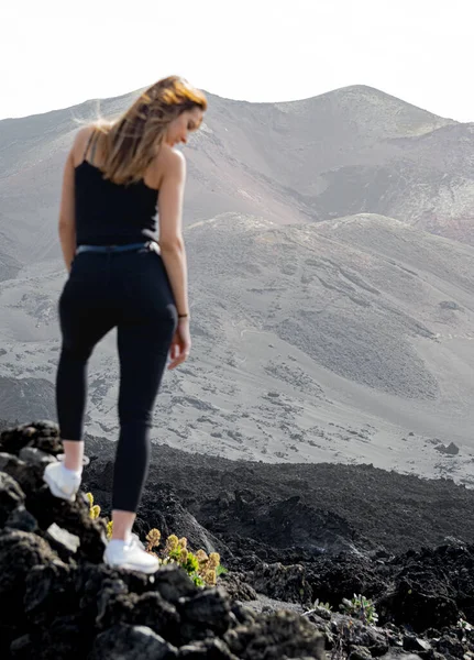 woman in suspenders standing with her hair flying in the wind looking at the ground in front of the Tajogaite volcano from a viewpoint, on top of some rocks, on the island of La Palma