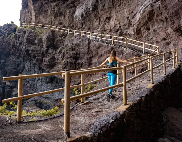 woman walking along a path between rocks on a cliff holding the railing with two hands, in La Palma, Canary Islands