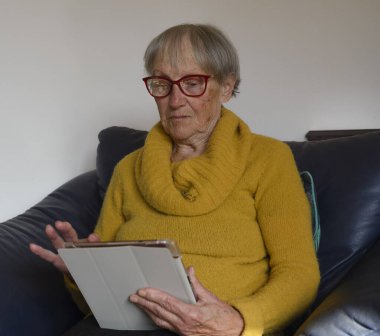 Bilgisayara bakarak dijital tablet kullanan olgun yaşlı bir kadın. Sanal aramalar, online toplantılar, televizyon izlemek, e-kitap okumak, teknolojik cihazlarla vakit geçirmek, evdeki kanepede oturmak.