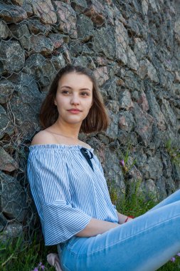Portrait of a young, Caucasian, short-haired woman, seated and leaning against a stone wall, enjoying the spring.