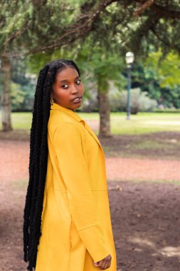 Saçlarında örgüler olan genç Afrikalı Amerikalı bir kadın, sırtına poz veriyor, arkasına bakıyor, sonbaharda sarı bir ceket giyiyor..