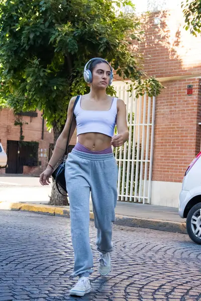 woman walks in the street in casual wear. She seems unaware if the photographer. She is wearing gray trousers, white sports a white crop-top and headphones. Behind the woman, the cityscape appears to be that of a nice neighborhood with calm streets