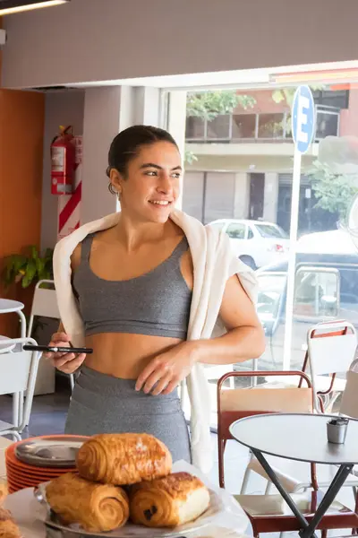 happy woman in a fitness bodysuit and fitness clothes ordering food through her cell phone in a bakery
