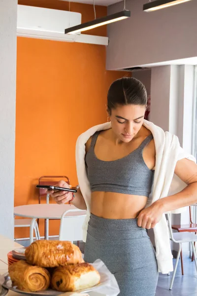 happy woman in a fitness bodysuit and fitness clothes ordering food on her cell phone in a bakery while arranging her clothing