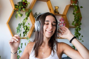 Genç siyah saçlı kadın evde boş zamanlarının tadını çıkarıyor. Oturma odasında kulaklıkla müzik dinliyor. Beyaz arka plan duvarı ve kahverengi süslemeleri var..