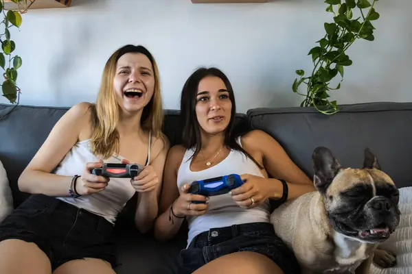 best friends playing video games on the couch at home having fun and laughing. young woman with fair complexion and young woman with tan complexion