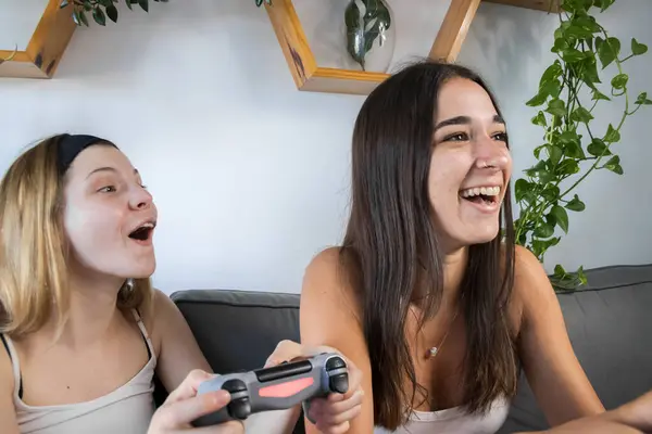 two friends laughing and playing video games. in the living room sitting on their couch. enjoying free time together. both wearing a white t-shirt and black shorts.