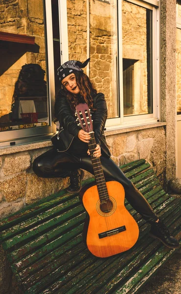 teenage woman with a guitar poured black with a headscarf perched on a wall on the side of a road