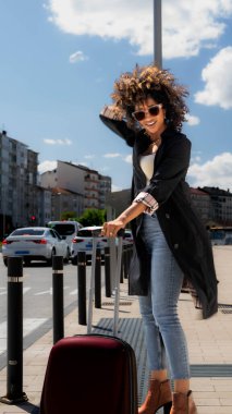 Güneş gözlüklü ve bavulu olan Afro-Amerikalı bir kadın.