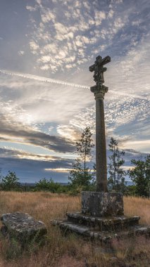Haç gökyüzüne yükseliyor, bir kaide üzerinde. Taşlar kuru bir çimenlikte, bulutlu bir gökyüzünün altında.