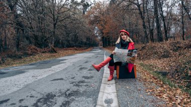 Bir bavul üzerinde oturan kadın yol kenarında açık bir kağıt haritayla bekliyor.