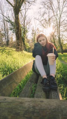 Genç bir kadın elinde bir bardakla parkta oturmuş samanla içiyor.
