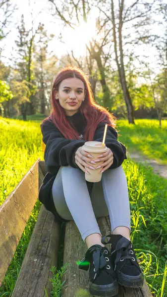 Genç bir kadın elinde bir bardakla parkta oturmuş samanla içiyor.