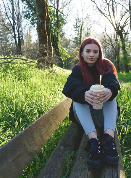 Genç bir kadın elinde bir bardakla parkta oturmuş samanla içiyor.