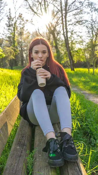 Genç bir kadın elinde bir bardakla parkta oturmuş samanla içiyor.