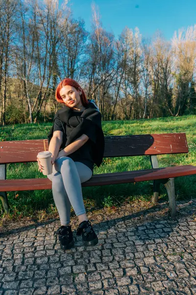 Genç bir kadın elinde bir bardakla parkta oturmuş samanla içiyor.