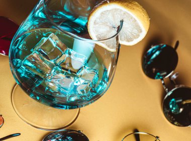 Bej arka planda, buzlu ve limon dilimli mavi bir kokteyl etrafında retro-stil güneş gözlüklü yaz kompozisyonu.