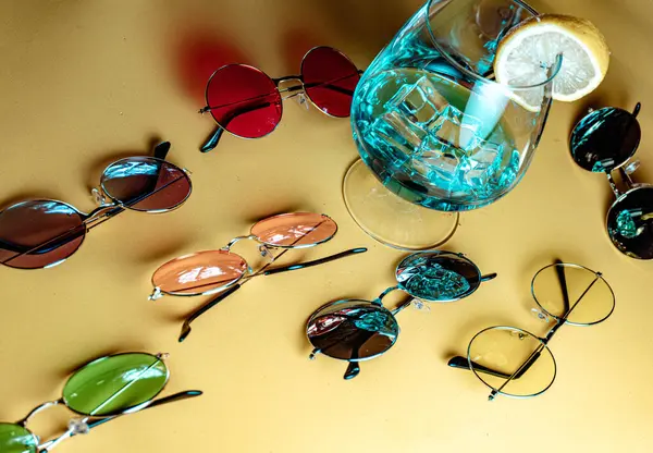 Bej arka planda, buzlu ve limon dilimli mavi bir kokteyl etrafında retro-stil güneş gözlüklü yaz kompozisyonu.