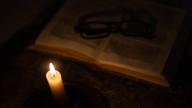 Işıklı bir mum, arka planda açık bir kitap ve odak dışı gözlüklerle karanlığı sıcacık bir şekilde aydınlatır. Bu da samimi ve sessiz bir okuma anını işaret eder..