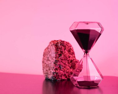 Elmas ve siyah kum şeklinde modern bir kum saati, desenli bir kayanın yanında, her ikisi de minimalist ve kavramsal bir atmosfer yaratan güçlü bir pembe ışıkla yıkanmış..