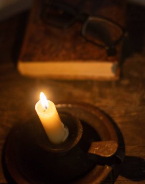 Işıklı bir mum, arka planda açık bir kitap ve odak dışı gözlüklerle karanlığı sıcacık bir şekilde aydınlatır. Bu da samimi ve sessiz bir okuma anını işaret eder..