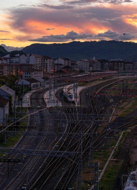 Ufuk çizgisine doğru uzanan geniş bir tren ağı beyaz bir trenin durmasıyla birlikte durdu. Arka planda, canlı bir günbatımı gökyüzünün altında binalar ve dağlar silueti var..