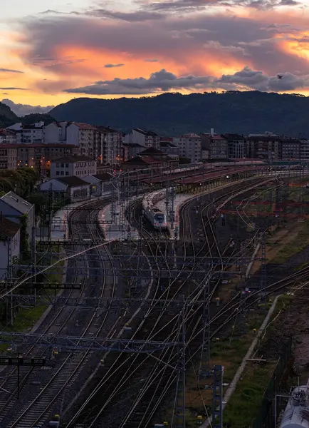 Ufuk çizgisine doğru uzanan geniş bir tren ağı beyaz bir trenin durmasıyla birlikte durdu. Arka planda, canlı bir günbatımı gökyüzünün altında binalar ve dağlar silueti var..