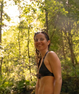 Bir kadın gülümsüyor ve kameraya bakıyor, saçları ıslak, aydınlık bir ormanda. Sahne mutluluk, tazelik ve doğal çevreyle derin bir bağ yayıyor..