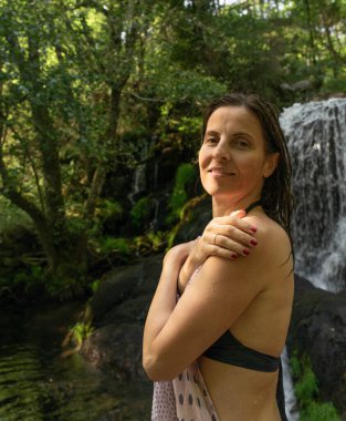 Omzunda eşarp olan bikinili bir kadın ormanlık alanda doğal bir şelalenin yanında serinliyor. Sakin ifadesi ve yeşil atmosferi, refahı ve doğayla olan bağlantısını iletir..