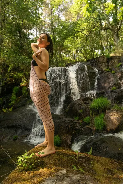 Kolu havada, başı arkada, kayanın üzerinde duran bir kadın yemyeşil bir ormanda doğal bir şelalenin tazeliğinin tadını çıkarıyor..