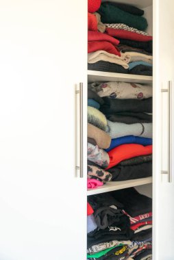 Bir dolap ya da soyunma odasının detaylı bir görüntüsü, askılarda asılı ve raflarda katlanmış çeşitli kıyafetleri gösteriyor. Görüntü, organizasyon ve geniş bir kostüm koleksiyonu öneriyor..