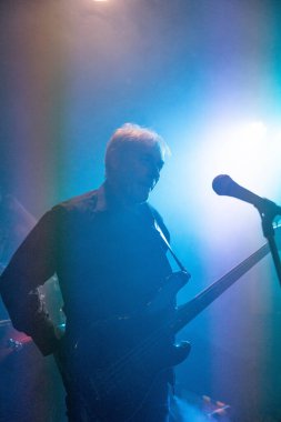 Bir müzisyenin silueti, muhtemelen bir basçı, sahnede, bir mikrofon ve enstrümanla, mavi konser ışıklarıyla aydınlatılmış. Görüntü canlı bir performans ve müzikal atmosferi çağrıştırıyor.