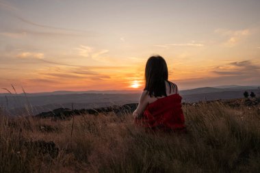 Siluet Sırtı dönük bir kadın, bir tepenin uzun çimlerinde oturan, güzel bir gün batımını izliyor. Güneş ufukta beliriyor, gökyüzünü altın ve turuncu renklerle boyuyor