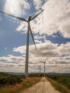 Büyük bir rüzgar türbininin geniş açılı görüntüsü, perspektifi düşük olan ve onun boyutunu ve varlığını vurgulayan perspektifi dönüyor. Bir toprak yol ufka doğru esiyor.